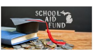 School Aid Fund written on chalk on a blackboard in front of a table with a graduation hat, a book and a pile of money to represent the Michigan fund receiving $1.3 Billion from the Michigan Lottery In 2023