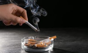 a hand puts a lit cigarette toward an ash tray full of cigarette butts